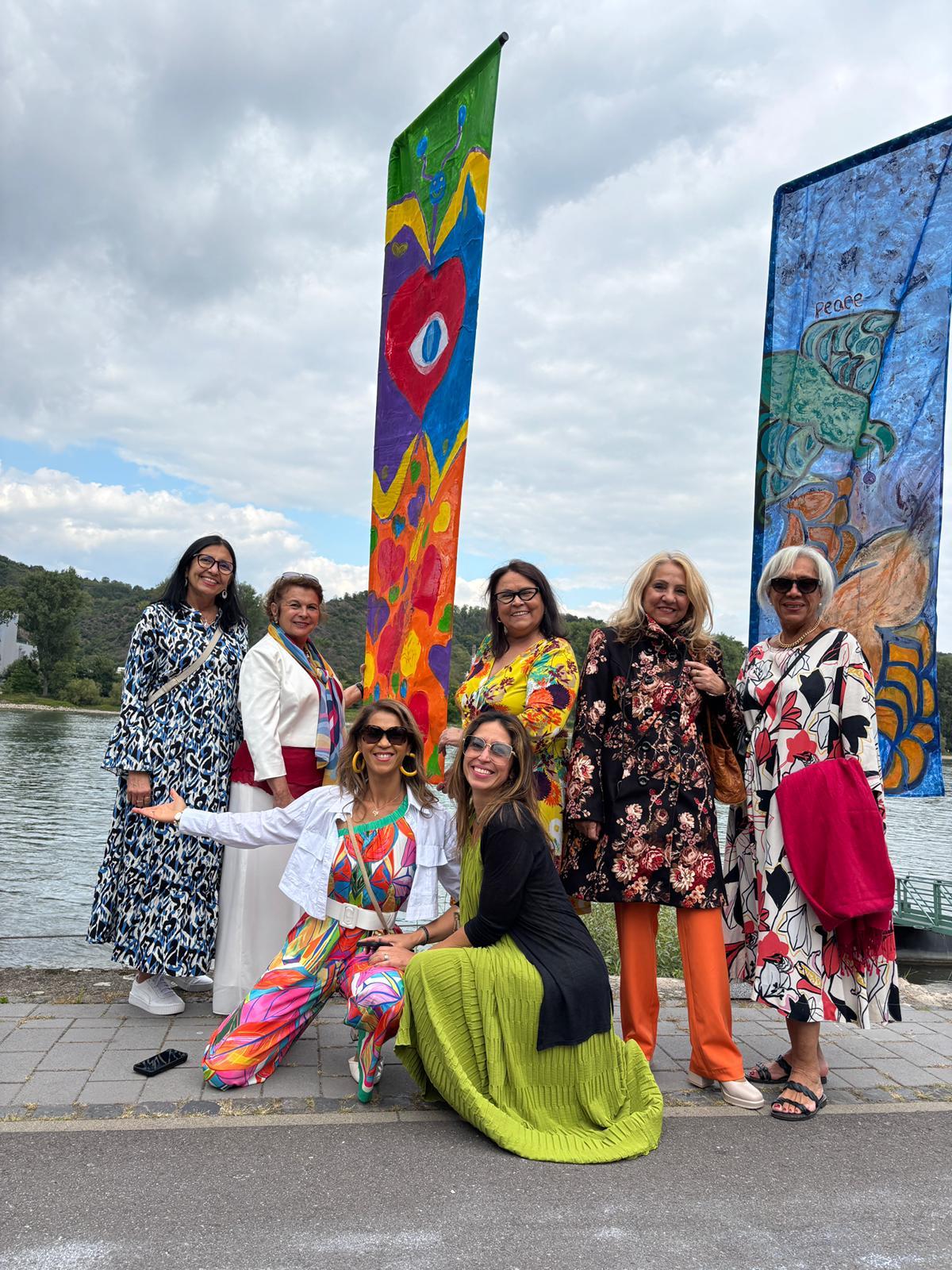 Um grupo de sete mulheres sorrindo, posando em frente a duas bandeiras coloridas de arte abstrata ao ar livre, perto de um rio. As mulheres vestem roupas vibrantes e estampadas.