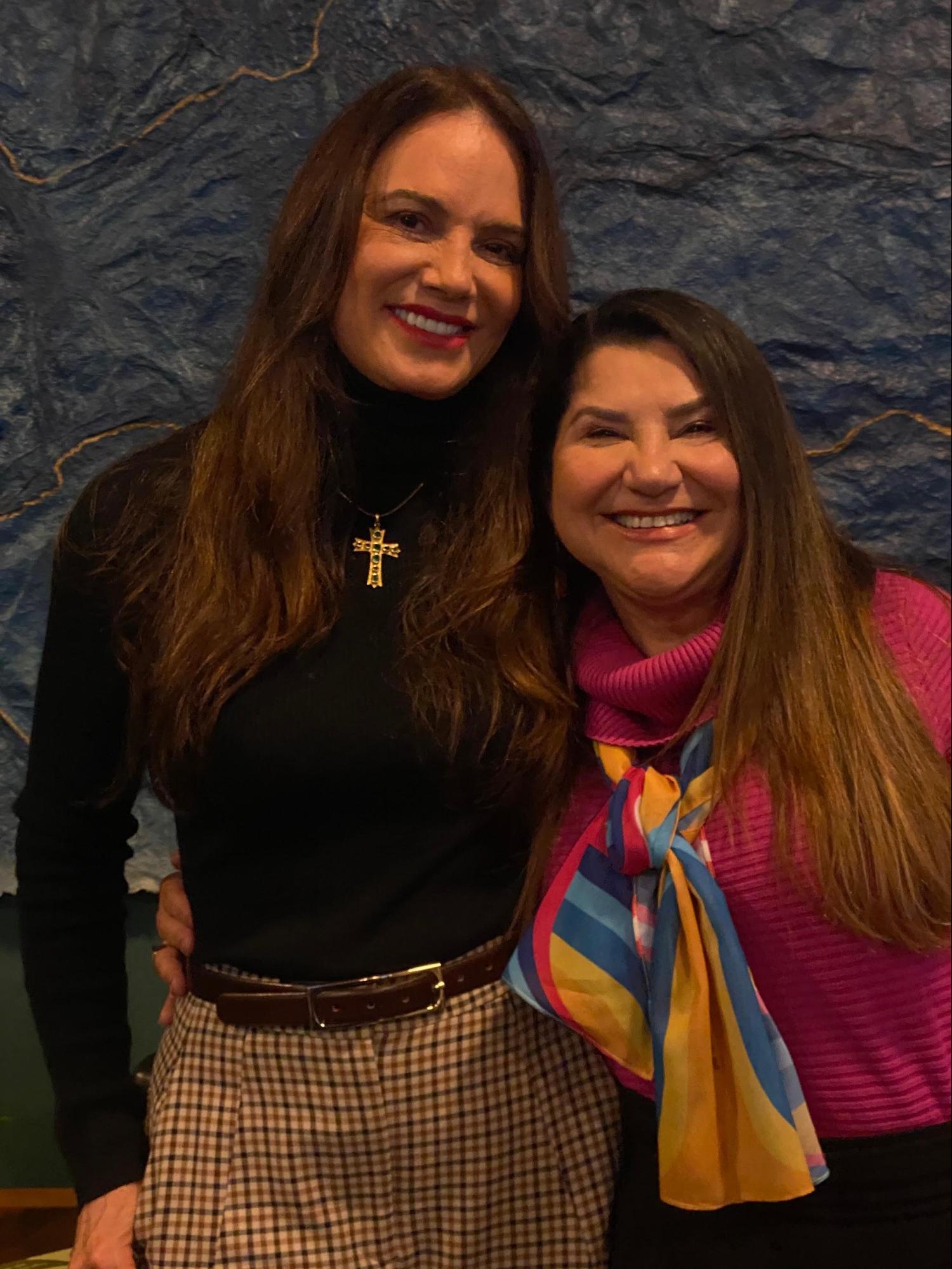 Duas mulheres sorridentes posando lado a lado. Uma veste uma blusa de gola alta preta e a outra veste um suéter rosa com um lenço colorido.