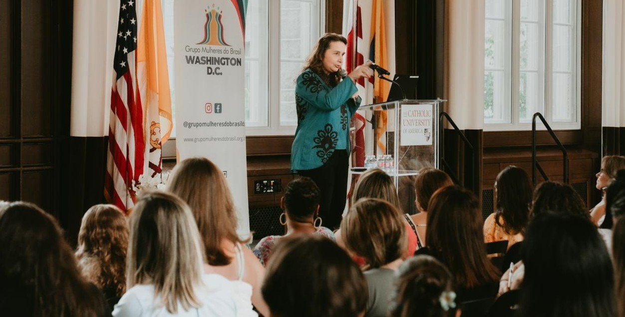 Uma mulher de pé no palco, falando e gesticulando em um microfone, em frente a um púlpito. Há um público composto por várias pessoas, e bandeiras e banners decoram o palco.
