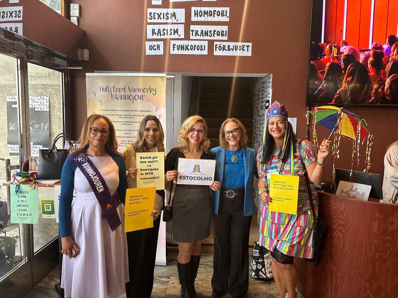 Cinco mulheres posando lado a lado em um saguão interno. Uma delas usa um traje festivo colorido e segura um pequeno guarda-chuva arco-íris. O fundo tem textos em sueco e uma tela.