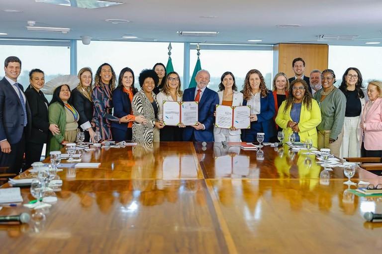 Um grupo de cerca de 20 pessoas, incluindo homens e mulheres, em torno de uma grande mesa de reunião de madeira brilhante. O presidente Lula está ao centro, com uma mulher ao lado, e ambos seguram documentos. Há duas bandeiras do Brasil ao fundo.