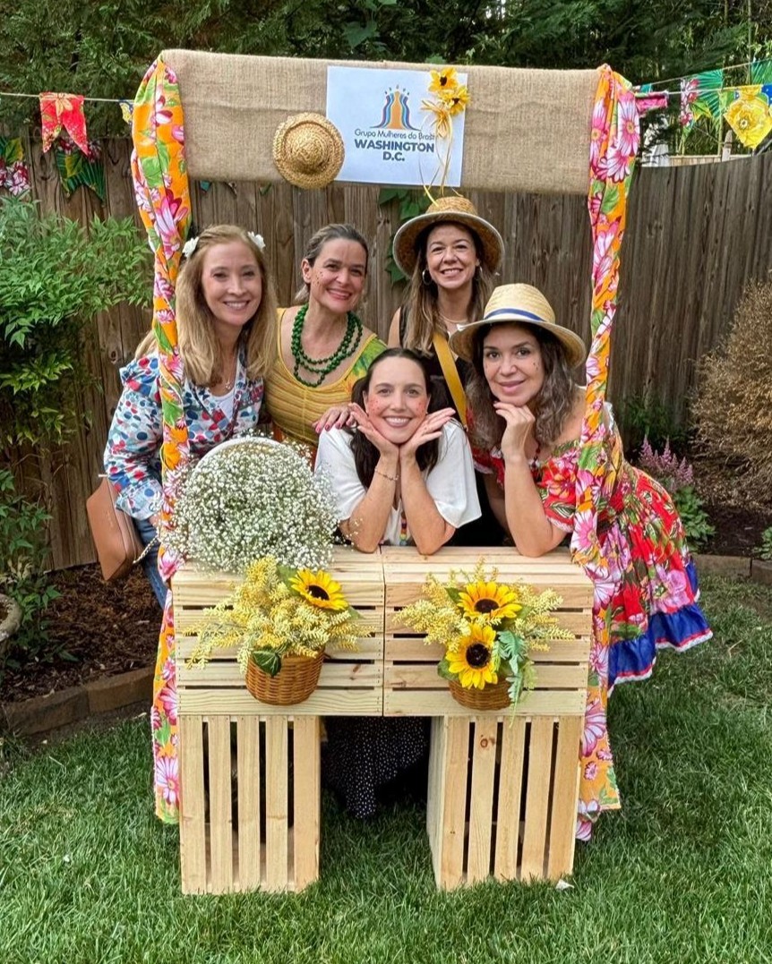 Cinco mulheres posando alegremente em uma barraca de madeira decorada para uma festa junina, com flores e bandeirinhas, em um jardim gramado.