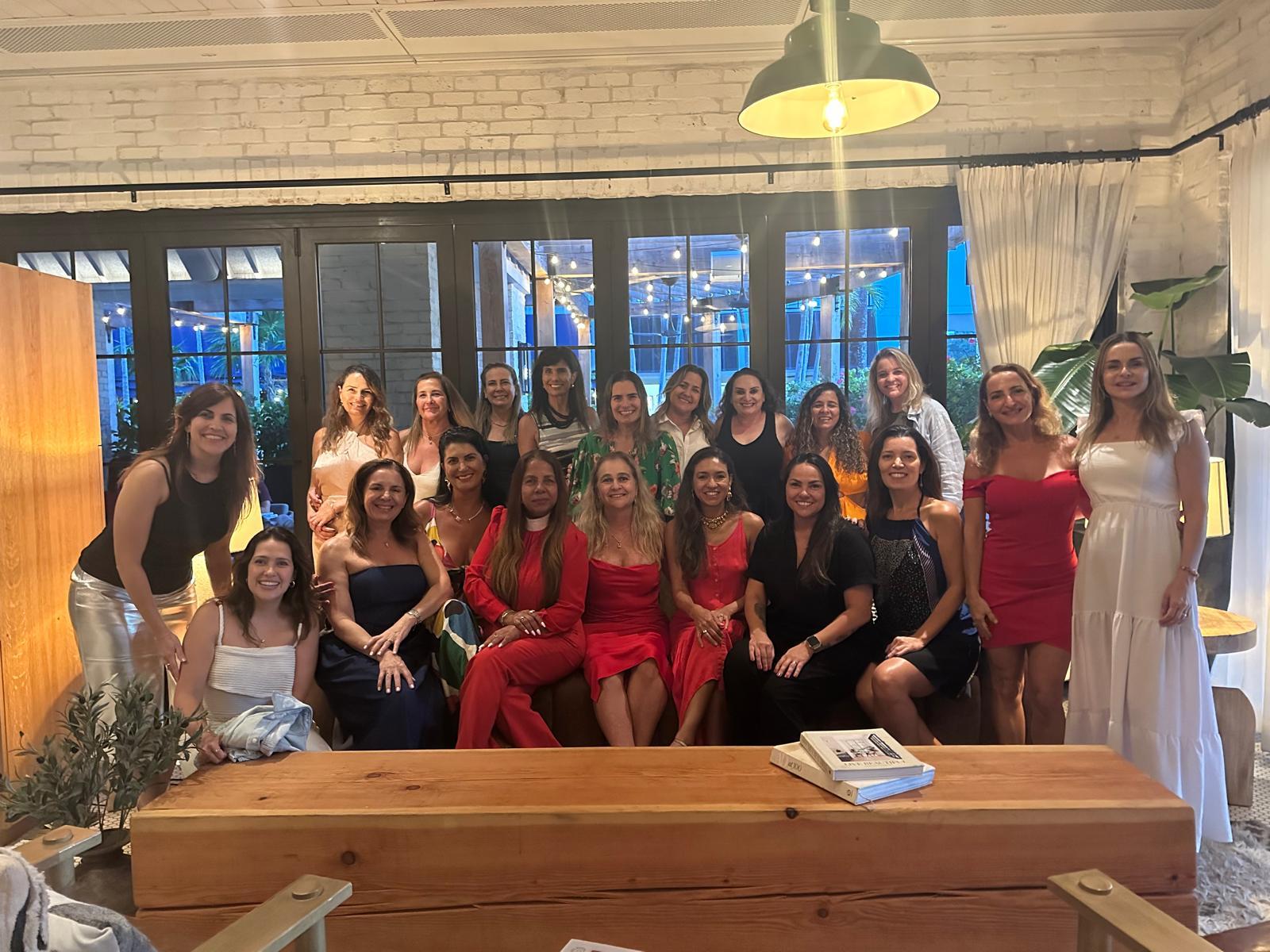 Um grupo de aproximadamente 20 mulheres em trajes elegantes (principalmente vestidos vermelhos, pretos e brancos) posando para uma foto em um sofá e atrás dele, em uma sala interna com grandes janelas ao fundo.