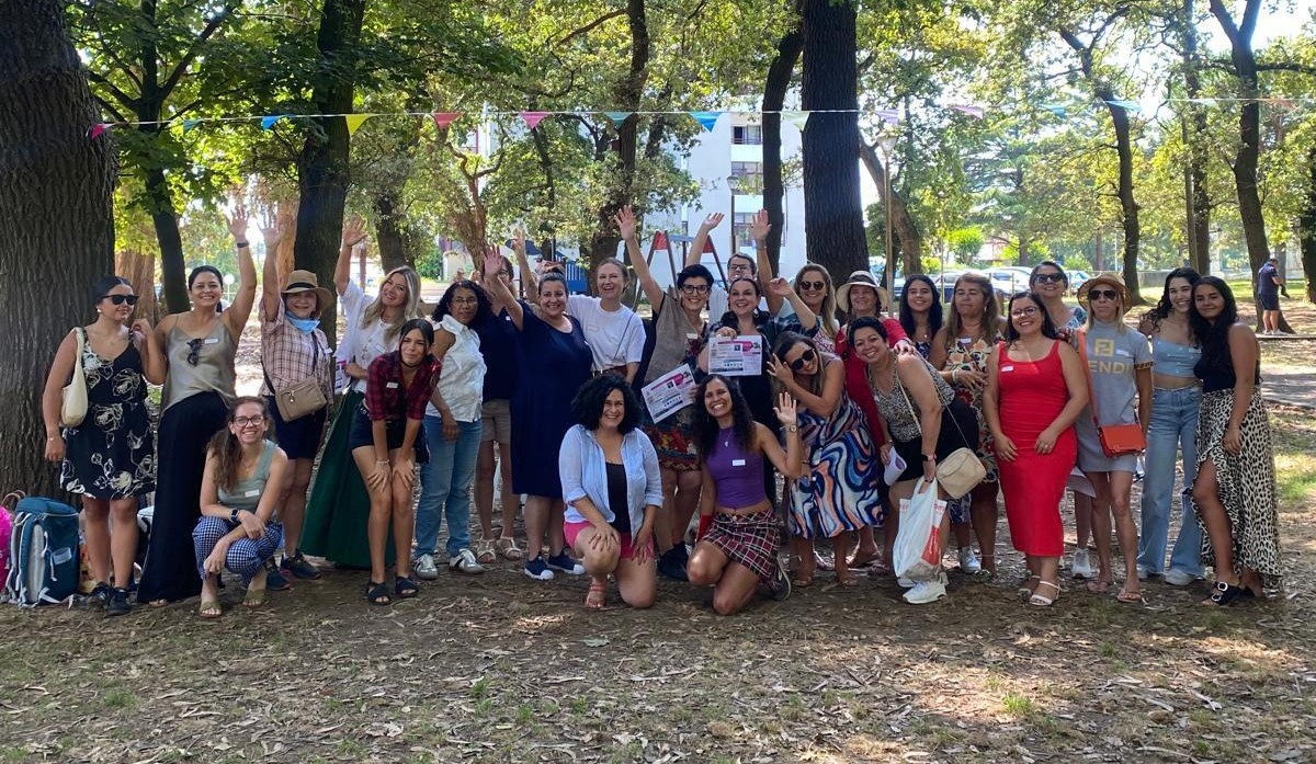 Um grande grupo de mais de 20 mulheres animadas, algumas de pé e outras agachadas, acenando para a câmera em um parque arborizado, sob a luz do sol.