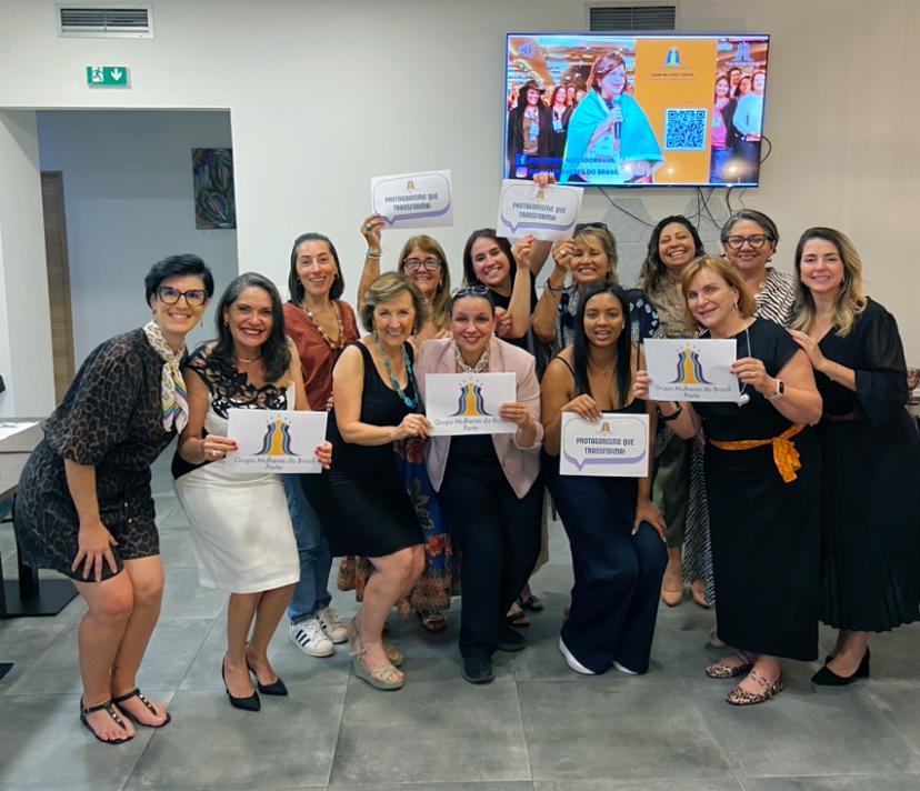 Um grupo de cerca de 13 mulheres sorrindo em um ambiente interno, a maioria segurando cartazes brancos com os dizeres “PROTAGONISMO QUE TRANSFORMA”. Uma tela de TV ao fundo exibe uma mulher em destaque.