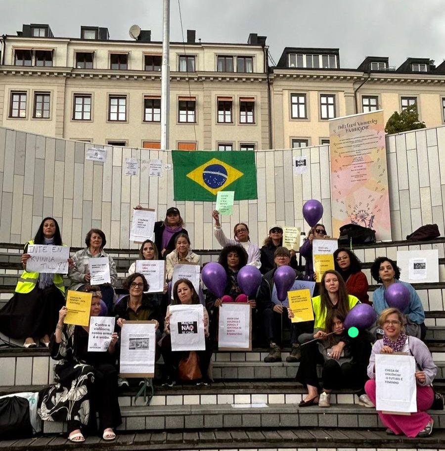 Um grupo de aproximadamente 15 mulheres em uma escadaria de rua, segurando cartazes e balões roxos. Uma bandeira do Brasil está pendurada ao fundo.
