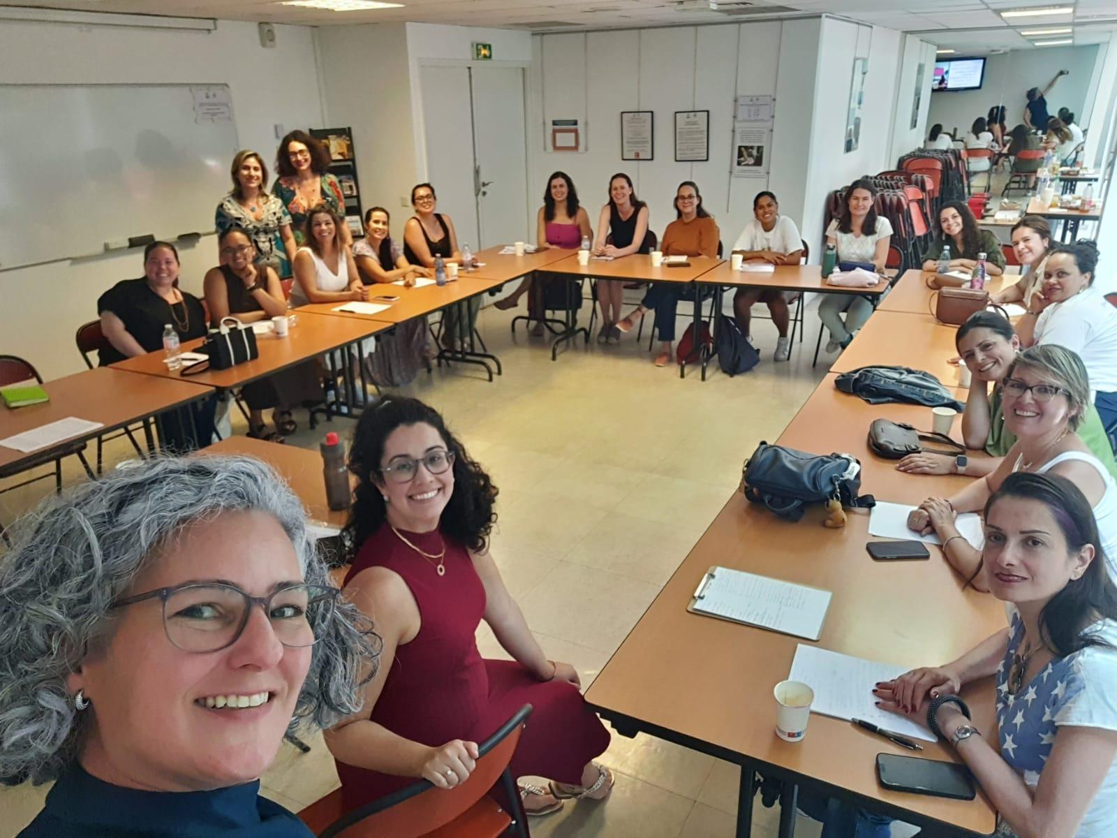 Uma selfie de grupo de cerca de 20 mulheres sentadas ao redor de mesas dispostas em formato de “U” em uma sala de aula. Todas estão sorrindo e olhando para a câmera.