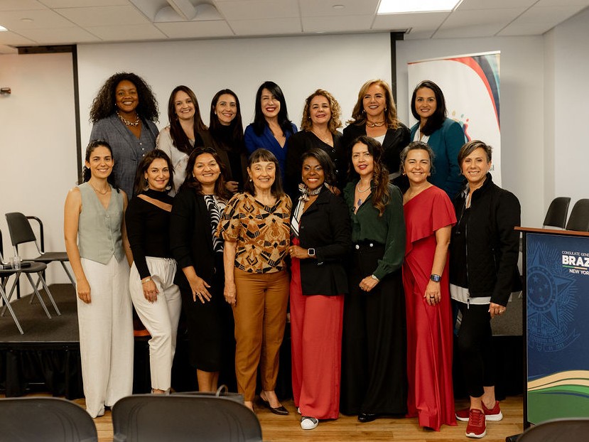 Cerca de quinze mulheres posam sorridentes em duas fileiras sobre um palco baixo. Elas vestem trajes casuais e formais em cores variadas como azul, vermelho, preto e tons neutros. Ao lado direito, vê-se um banner e um púlpito azul com o brasão do Brasil e a inscrição 'Consulate General of Brazil in New York'.
