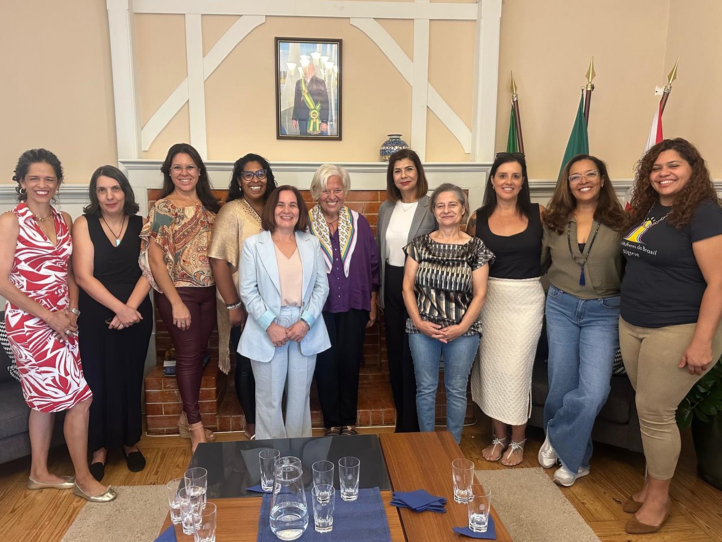Onze mulheres sorridentes posam em pé em uma sala de estar com piso de madeira. Ao centro, atrás delas, há uma lareira de tijolos e, acima dela, o retrato oficial do Presidente da República. No lado direito do ambiente, estão posicionadas as bandeiras do Brasil e de Portugal.