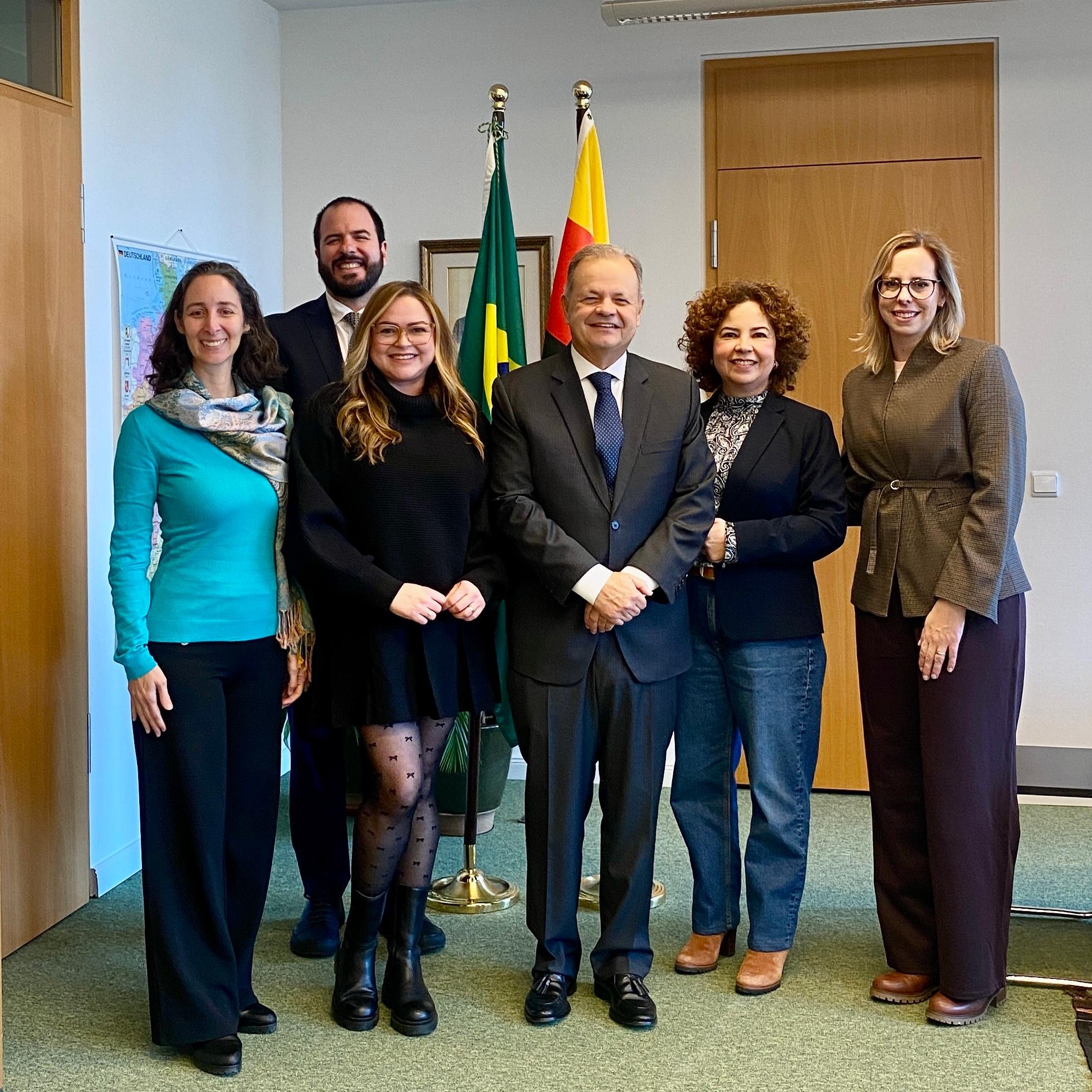 Cinco mulheres e dois homens posam sorridentes em um escritório com as bandeiras do Brasil e da Alemanha ao fundo. Eles vestem trajes formais e casuais de inverno, como blazers, suéteres e cachecóis. À esquerda, um mapa está fixado na parede branca.