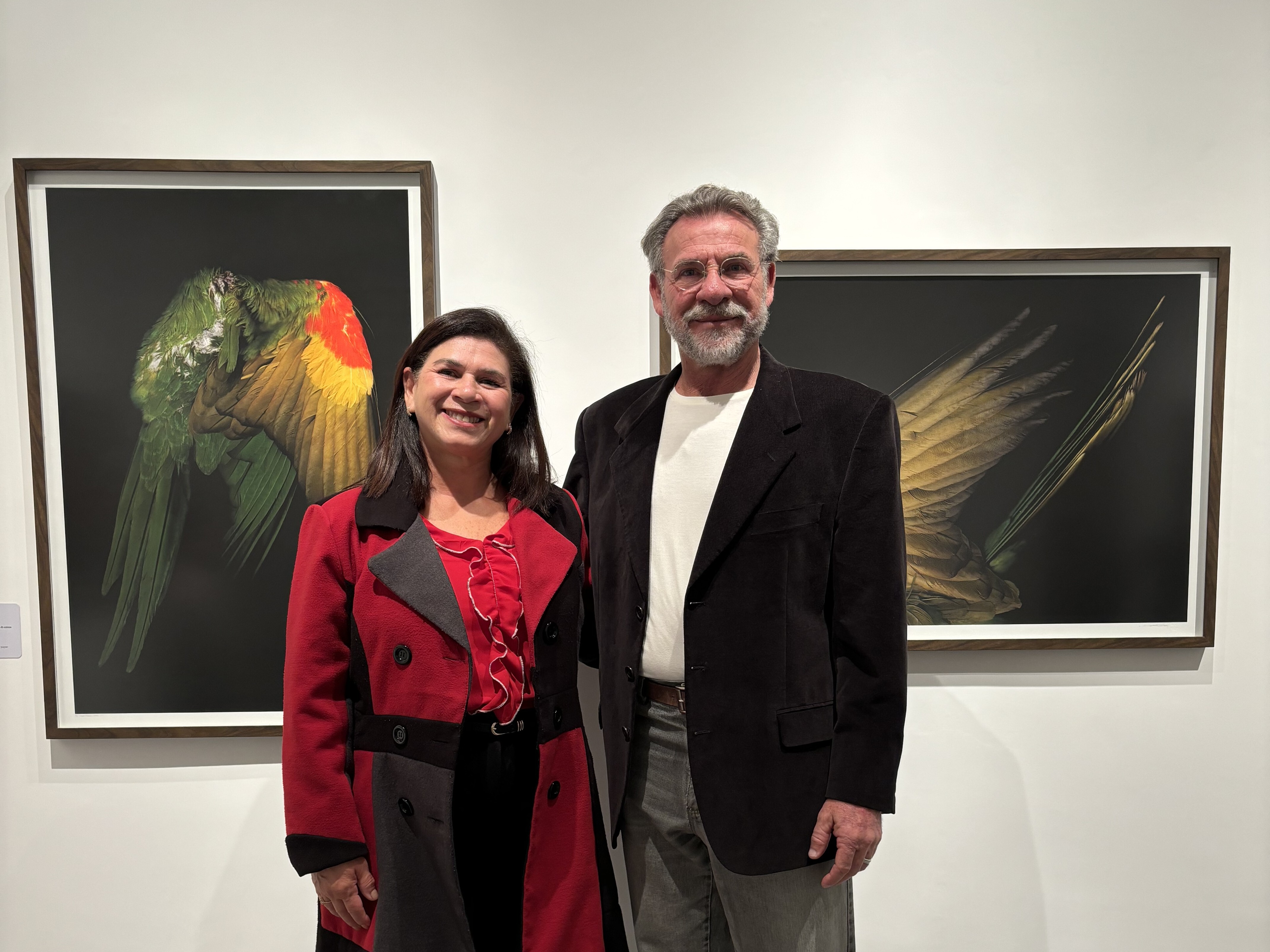 Uma mulher e um homem posam sorridentes em uma galeria de arte de paredes brancas. A mulher veste um casaco vermelho com lapelas cinzas sobre uma blusa de babados, e o homem veste um paletó escuro com camisa branca. Ao fundo, estão expostas duas fotografias emolduradas em madeira: à esquerda, a imagem detalhada de uma ave verde e amarela; à direita, o detalhe de penas em tons terrosos sobre um fundo preto.