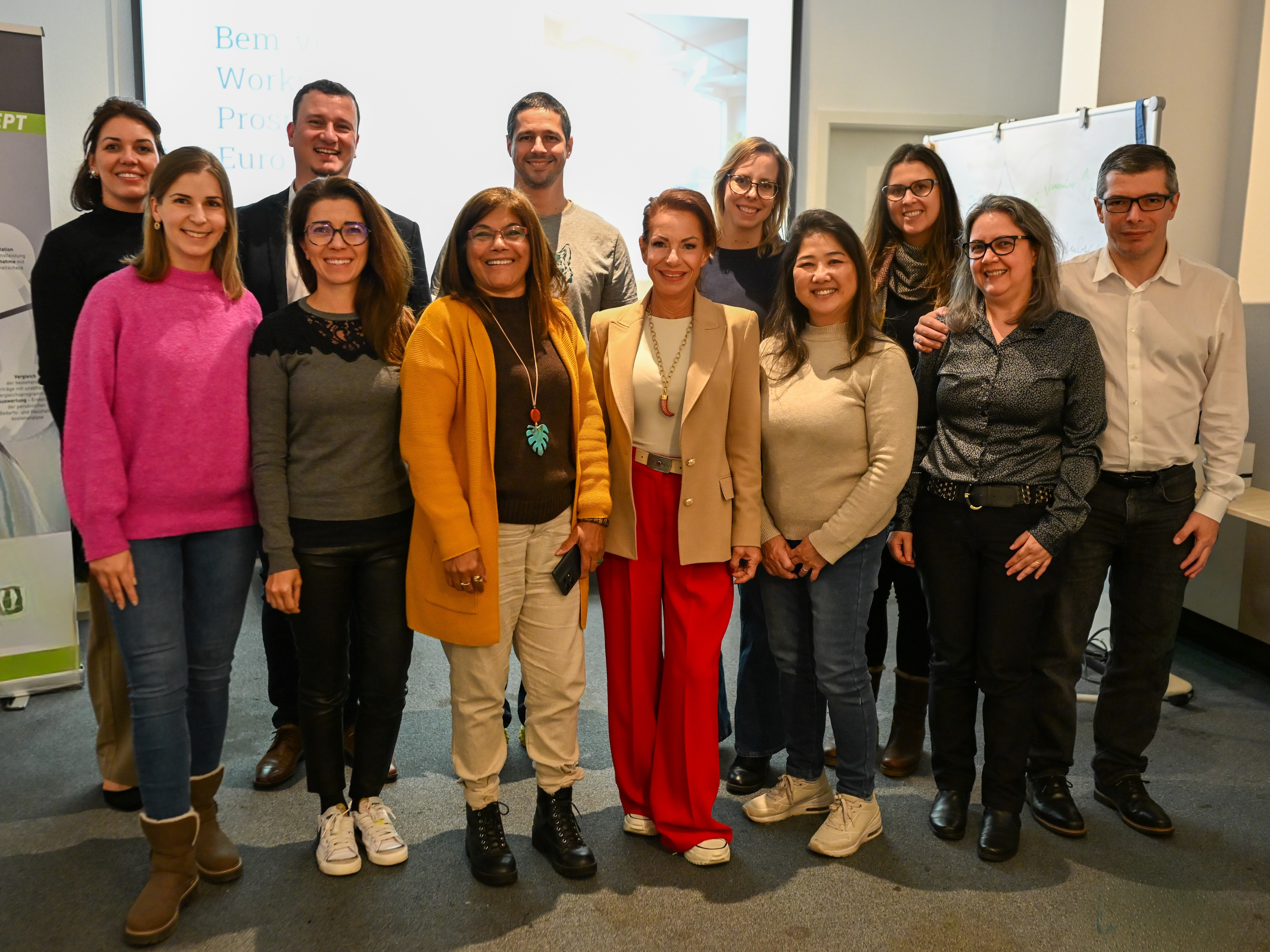Um grupo de doze pessoas, sendo nove mulheres e três homens, posam sorridentes para a foto em uma sala de treinamento. Elas vestem roupas casuais e sociais em cores variadas, com destaque para uma mulher ao centro com calças vermelhas vibrantes e blazer bege. Ao fundo, vê-se uma tela de projeção com o título do workshop e um quadro branco à direita com anotações.