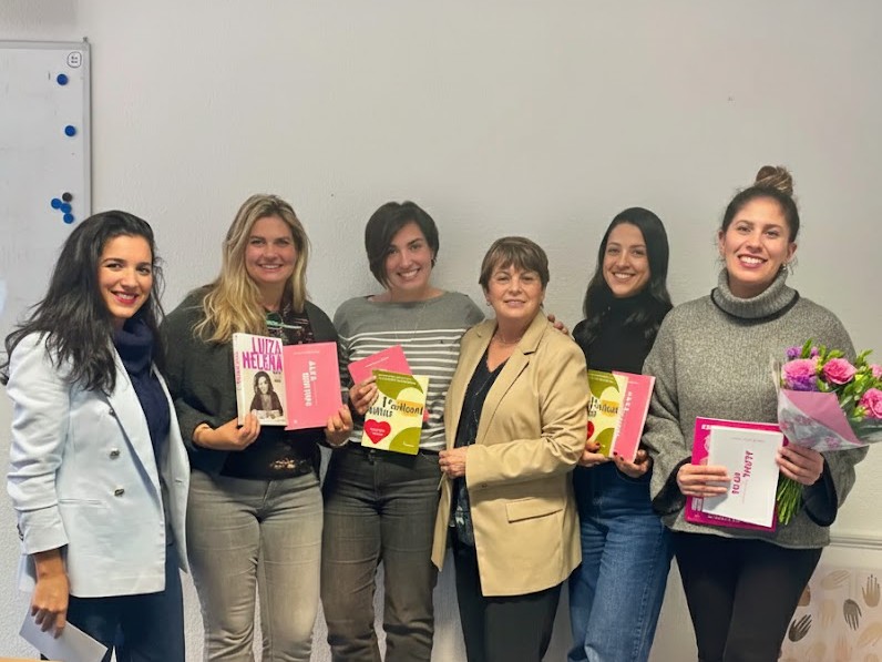Seis mulheres sorridentes posam lado a lado em frente a uma parede branca. Elas seguram livros com capas coloridas em tons de rosa, amarelo e branco, e a mulher à extrema direita segura também um buquê de flores em tons de rosa e roxo. Elas vestem roupas casuais e formais, como blazers, blusas de tricô e jeans. À esquerda, parte de um quadro branco com ímãs coloridos é visível.