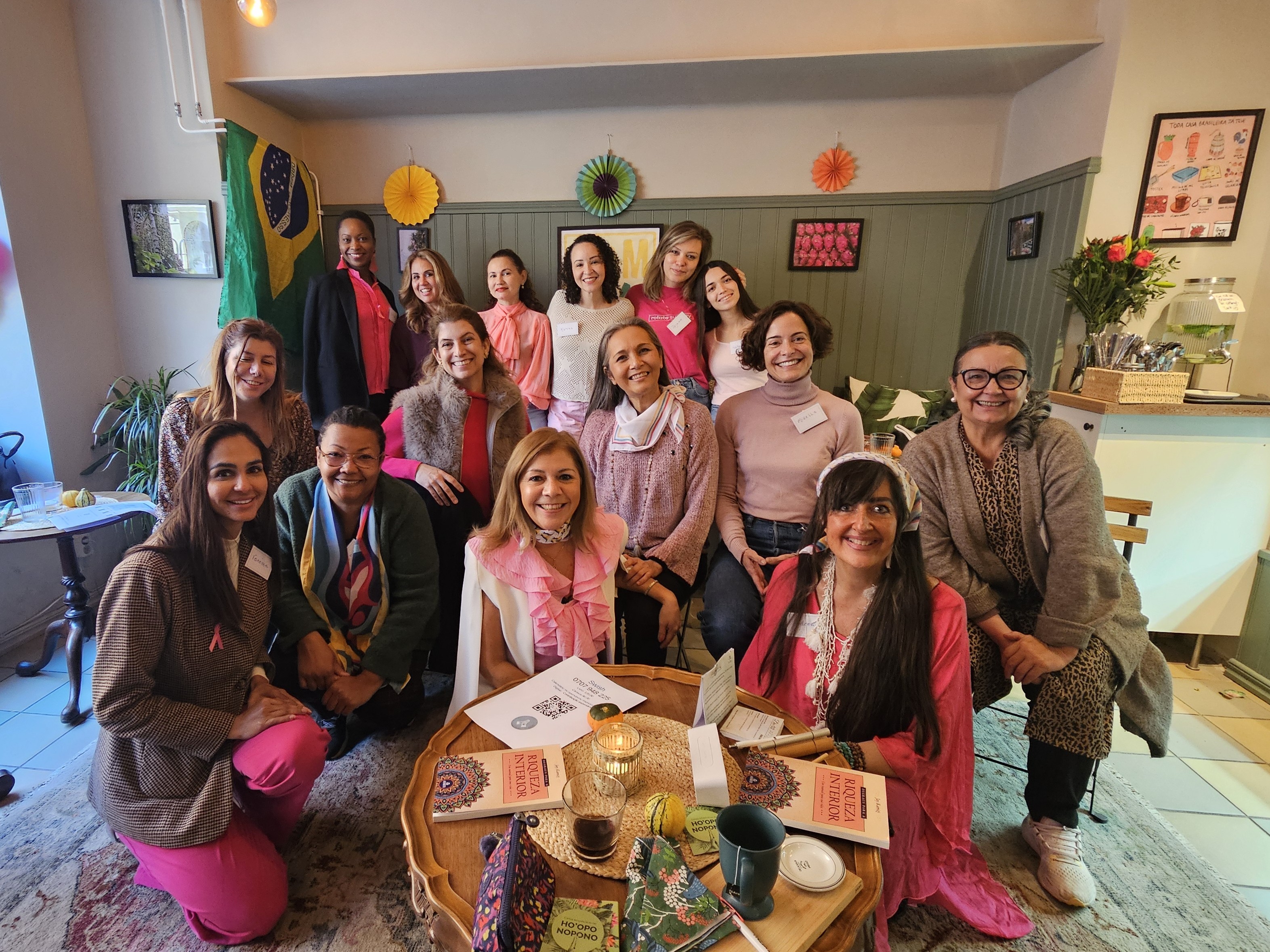 Um grupo de dezesseis mulheres sorri em um ambiente acolhedor decorado com uma bandeira do Brasil ao fundo. Algumas estão sentadas ao redor de uma mesa de centro redonda que exibe livros intitulados 'Riqueza Interior', uma vela e objetos decorativos, enquanto as demais se posicionam em pé atrás delas. A maioria veste tons de rosa e bege. A sala possui paredes verdes com detalhes em madeira e pequenos enfeites circulares coloridos.