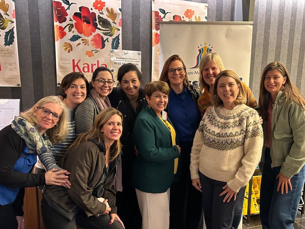 Nove mulheres sorridentes estão reunidas em um ambiente interno. Elas vestem roupas de frio, como casacos, blusas de lã com estampas geométricas e cachecóis. Ao fundo, as paredes cinza possuem listras verticais e estão decoradas com cartazes artísticos de flores vermelhas e a palavra 'Karla'. À direita, um banner branco exibe o logotipo colorido do Grupo Mulheres do Brasil.