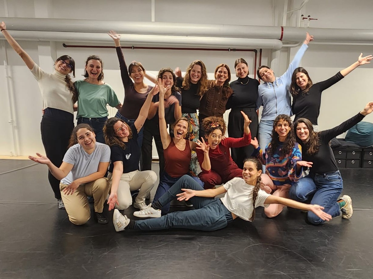 Um grupo de dezessete mulheres celebra animadamente em um estúdio de dança com piso preto e paredes brancas. Elas estão distribuídas em diferentes níveis: algumas em pé ao fundo com os braços erguidos, outras agachadas ao centro e uma mulher deitada de lado à frente com os braços abertos. Elas vestem roupas confortáveis e esportivas, como camisetas, leggings e calças de moletom, exibindo expressões de alegria e descontração.