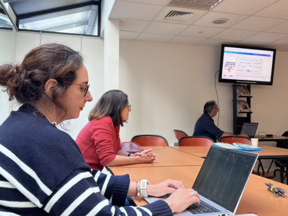 Em um ângulo lateral, duas mulheres estão sentadas em uma mesa comprida de madeira em uma sala de aula. A mulher em primeiro plano tem cabelos presos, usa óculos e veste um suéter listrado de azul e branco enquanto digita em um notebook prateado. Ao fundo, uma terceira pessoa trabalha em outro computador sob uma tela de TV fixada na parede, que exibe slides de uma apresentação. O ambiente é iluminado por uma claraboia no teto.