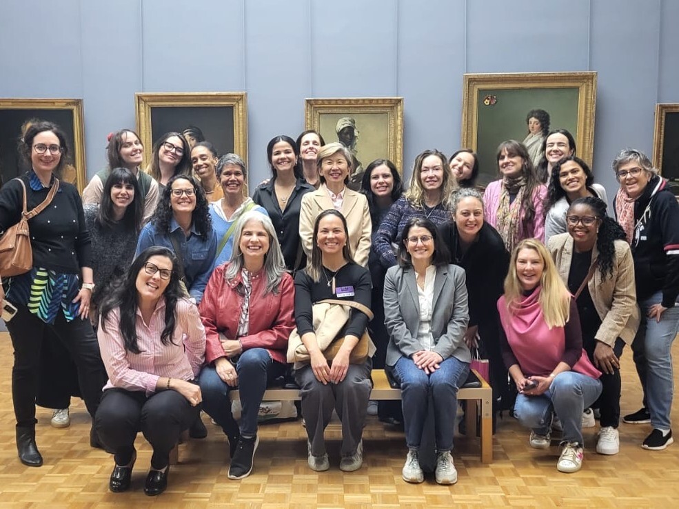 Um grupo de vinte e cinco mulheres de diferentes idades posa sorridente em uma galeria de arte com paredes azuis acinzentadas. Elas estão organizadas em três fileiras, com algumas sentadas em um banco central e as demais em pé ou agachadas ao redor. Ao fundo, estão expostas diversas pinturas clássicas em molduras douradas, retratando figuras humanas. O ambiente possui piso de madeira clara em padrão de blocos.