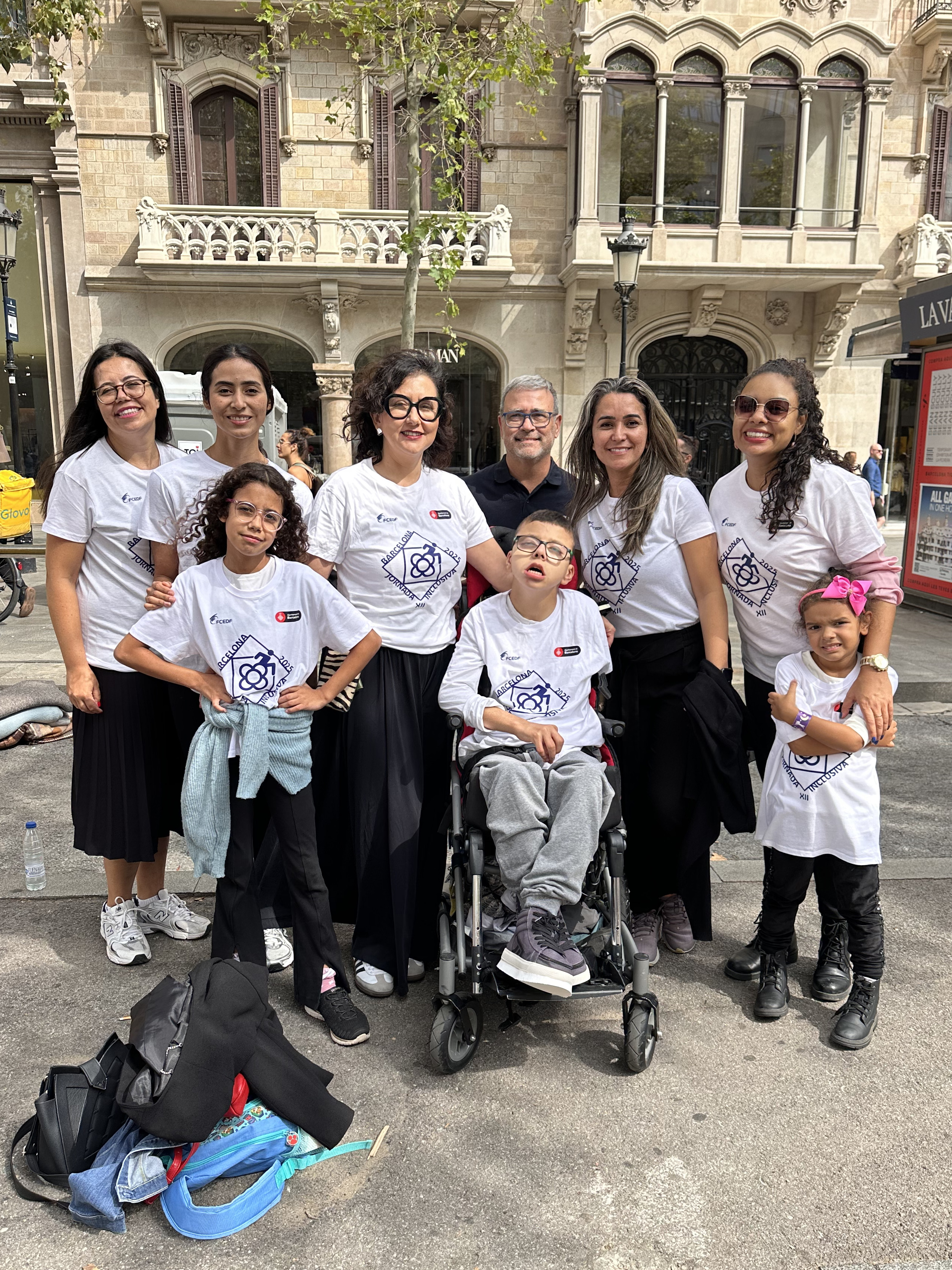 Um grupo de dez mulheres posa sorridente em um ambiente externo pavimentado, em frente a um prédio moderno com fachada de vidro. Algumas participantes seguram uma grande bandeira branca com o logotipo colorido do Grupo Mulheres do Brasil, enquanto outras sustentam uma moldura de papelão que simula uma postagem de rede social com a frase 'Dia de Inclusão e Esporte'. Elas vestem roupas casuais e esportivas, e o reflexo do céu nublado é visível nos vidros ao fundo.
