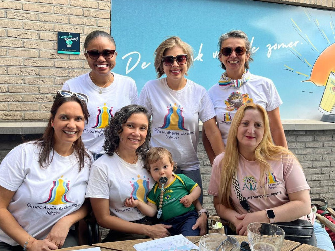 Cinco mulheres sorridentes e uma criança pequena posam juntas em uma área externa. Elas vestem camisetas brancas com o logotipo colorido do 'Grupo Mulheres do Brasil - Bélgica', enquanto a criança usa uma camisa verde da seleção brasileira. Três mulheres estão em pé ao fundo e duas sentadas à frente, ao redor de uma mesa de madeira.
