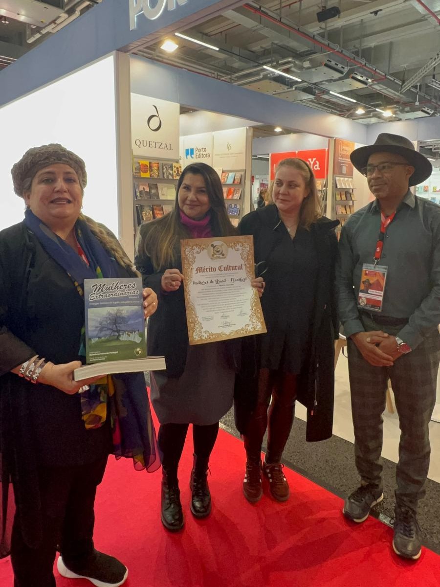 Três mulheres e um homem posam sobre um tapete vermelho em um estande da feira literária. No centro, uma mulher segura um certificado de moldura ornamentada intitulado 'Mérito Cultural'. À esquerda, outra mulher segura um livro com o título 'Mulheres Extraordinárias'. Todos sorriem para a câmera, com as prateleiras de livros ao fundo sob o letreiro 'Quetzal'.