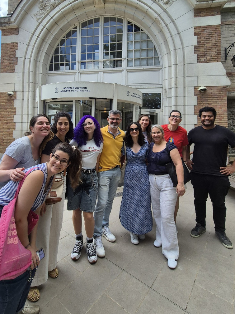 Grupo de dez pessoas, majoritariamente mulheres, posam sorridentes em frente à entrada arqueada do Hospital Fondation Adolphe de Rothschild. O grupo apresenta estilos variados, incluindo uma jovem com cabelo roxo vibrante e uma mulher com vestido longo azul estampado. Eles estão agrupados de forma descontraída na calçada clara, sob uma grande janela semicircular que compõe a fachada clássica do edifício.
