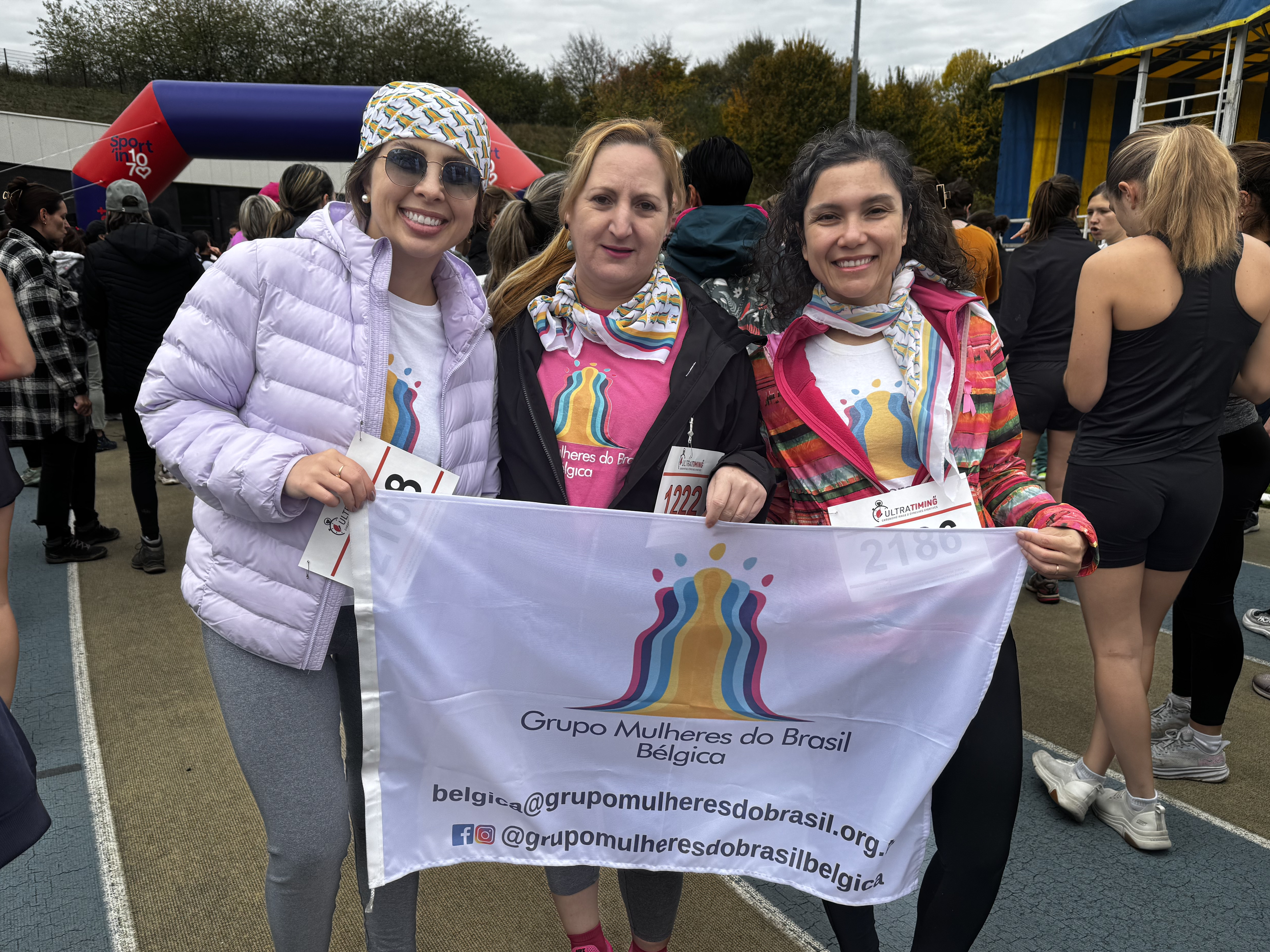 Três mulheres sorridentes posam em uma pista de atletismo ao ar livre, segurando juntas uma bandeira branca com o logotipo colorido do 'Grupo Mulheres do Brasil - Bélgica'. Elas vestem roupas esportivas e lenços estampados na cabeça ou no pescoço; a mulher à esquerda usa uma jaqueta acolchoada lilás, a do centro uma camiseta rosa e a da direita uma jaqueta colorida aberta. Ao fundo, vê-se um arco inflável de evento esportivo e outras pessoas participando da atividade.
