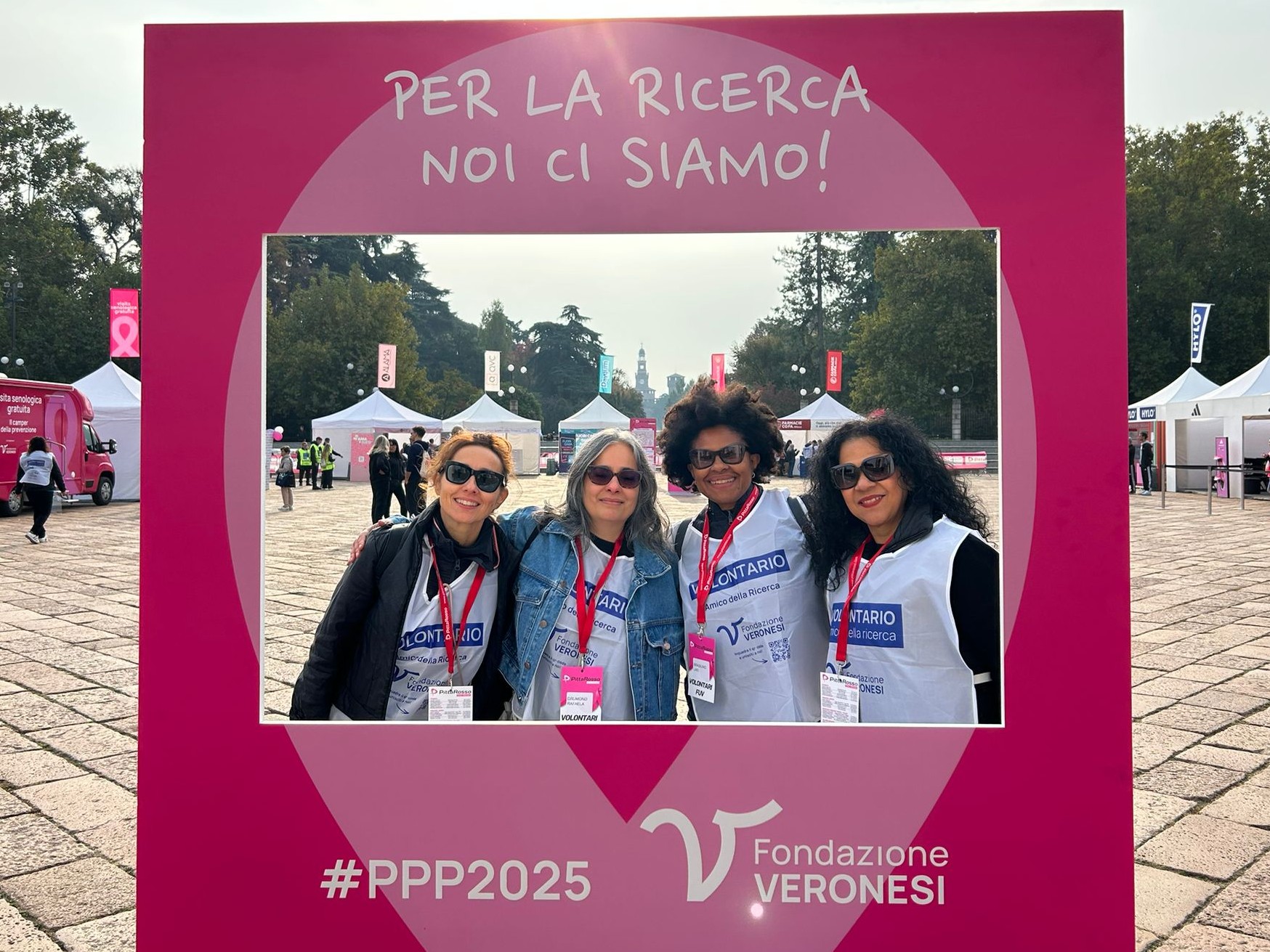 Quatro mulheres sorridentes posam juntas dentro de uma moldura fotográfica gigante de cor rosa vibrante. Elas usam coletes brancos com a palavra 'VOLONTARIO' e crachás pendurados no pescoço. A moldura contém o texto 'PER LA RICERCA NOI CI SIAMO!' e o logotipo da 'Fondazione Veronesi' na parte inferior. Ao fundo, vê-se uma praça ampla com tendas brancas de eventos e árvores sob um céu claro.
