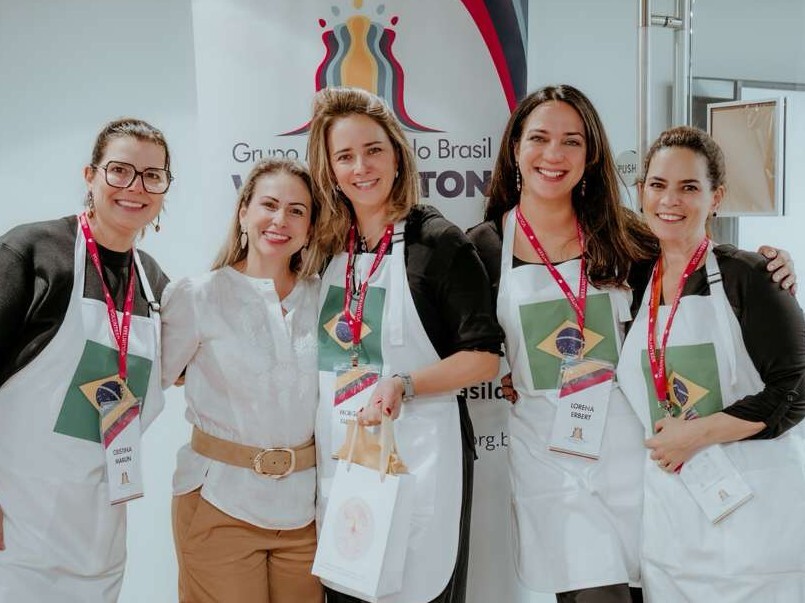 Cinco mulheres sorridentes posam juntas em um ambiente interno iluminado. Quatro delas vestem aventais brancos sobre roupas pretas, cada avental exibindo uma pequena bandeira do Brasil no centro e crachás com cordões vermelhos. A mulher ao centro, vestindo uma blusa branca e calça bege, segura uma sacola de papel branca. Ao fundo, uma faixa branca exibe parcialmente o logotipo colorido do Grupo Mulheres do Brasil.