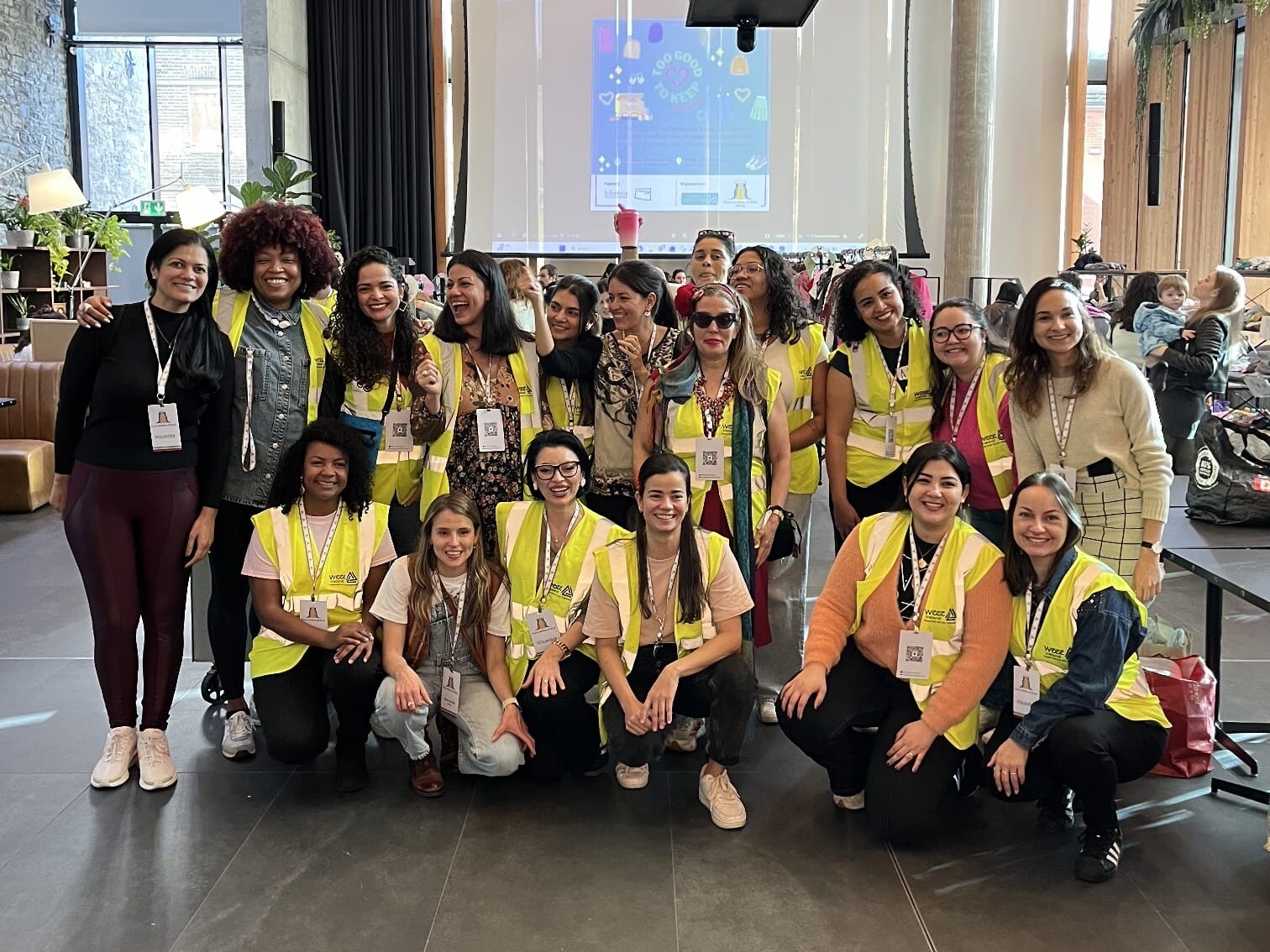 Um grande grupo de mulheres sorridentes posam em um espaço amplo e moderno com teto alto. A maioria veste coletes de segurança amarelos neon sobre suas roupas e exibe crachás brancos no pescoço. Oito mulheres estão em pé ao fundo, enquanto outras oito estão agachadas à frente. Ao fundo, uma projeção em uma tela grande mostra um cartaz azul do evento, e grandes janelas iluminam o ambiente.