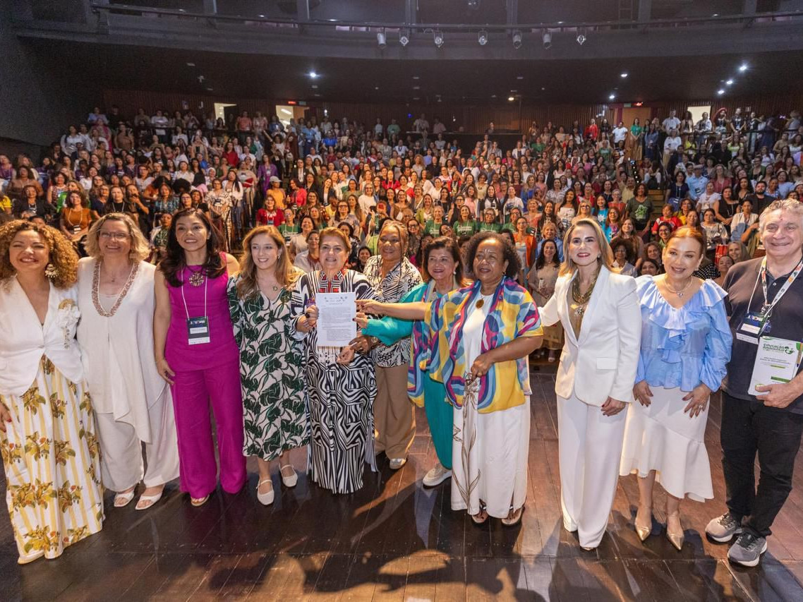 Uma ampla plateia composta majoritariamente por mulheres lotou o auditório com assentos em níveis elevados ao fundo. No palco, em primeiro plano, Luiza Helena Trajano e um grupo de doze pessoas posam para a foto. No centro, elas seguram juntas a Carta das Mulheres pelo Clima. O grupo veste trajes variados e elegantes, incluindo blazers brancos, vestidos com estampas botânicas e geométricas, e uma túnica colorida com ondas em tons de azul e amarelo.