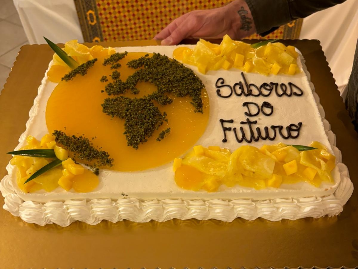 Bolo retangular decorado com cobertura branca e detalhes laterais em Chantilly. No topo, à esquerda, há uma representação do mapa-múndi feita com geleia amarela e granulado verde por cima. À direita, a frase 'Sabores do Futuro' está escrita em caligrafia preta. Pedaços de frutas amarelas e pequenas folhas verdes decoram as extremidades e o contorno do mapa.