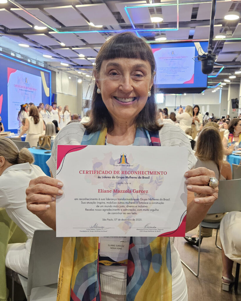 Mulher sorrindo e segurando um certificado de reconhecimento em um evento com público ao fundo.