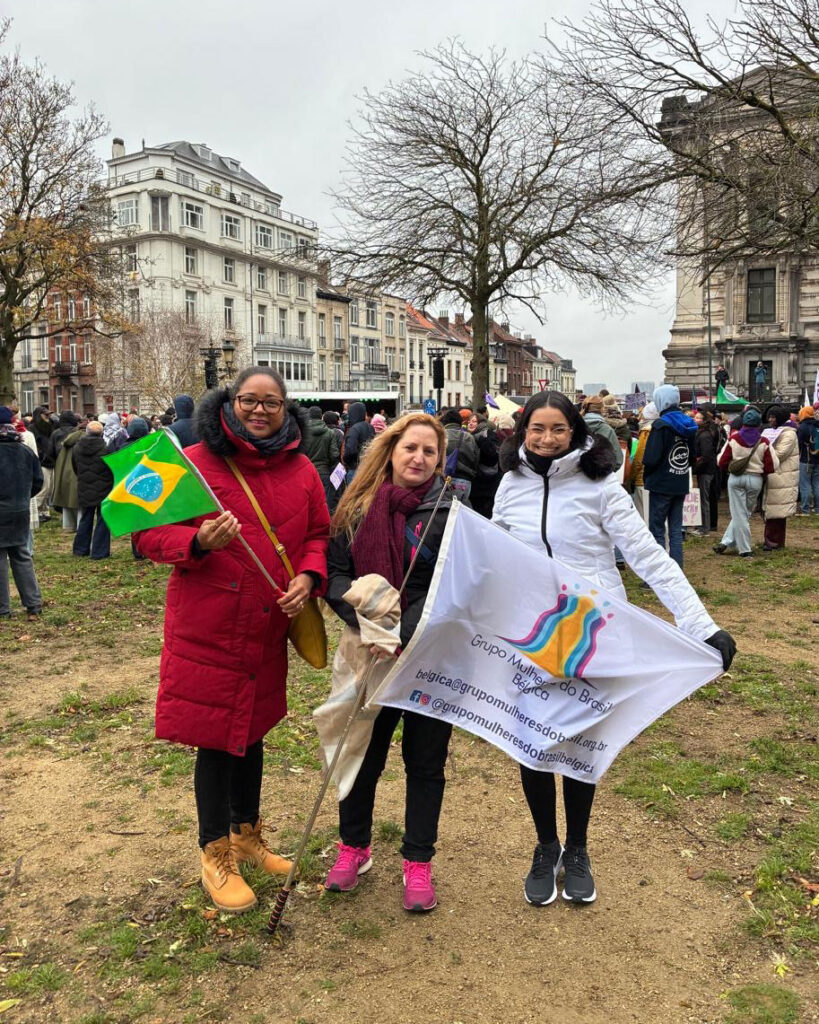 Três mulheres participando de uma manifestação ao ar livre segurando bandeiras do Brasil e do Grupo Mulheres do Brasil.