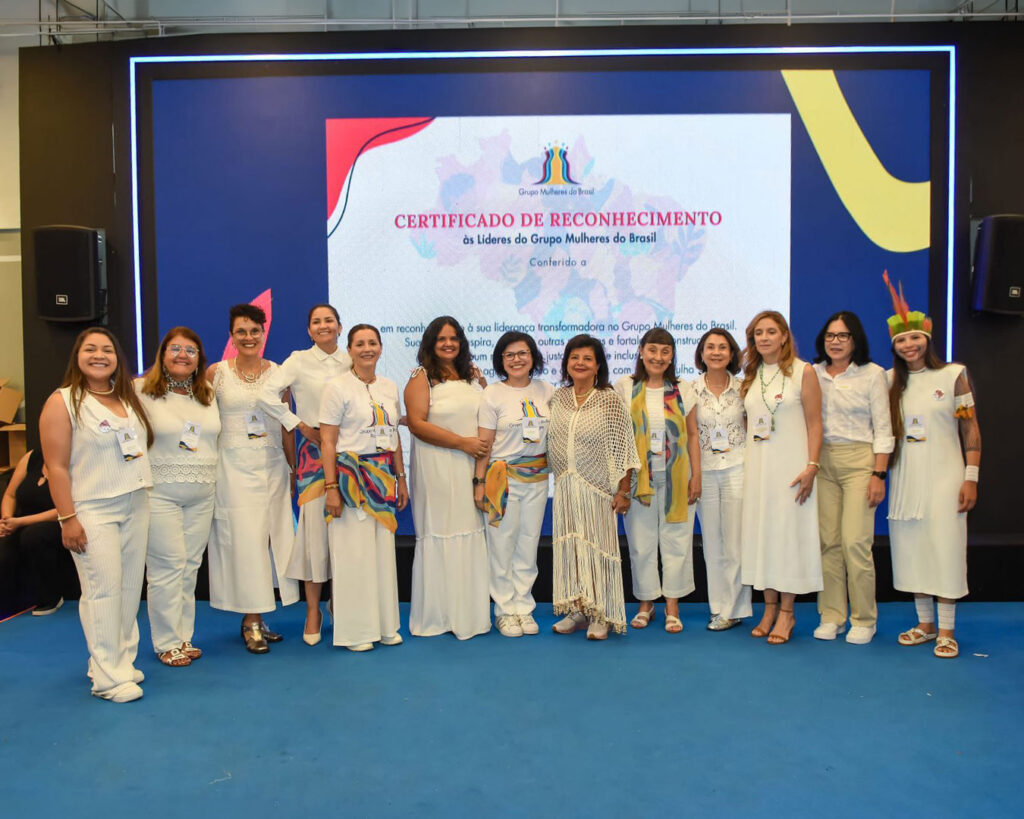 Grupo de mulheres vestidas de branco posando em um palco diante de um telão com certificado de reconhecimento.