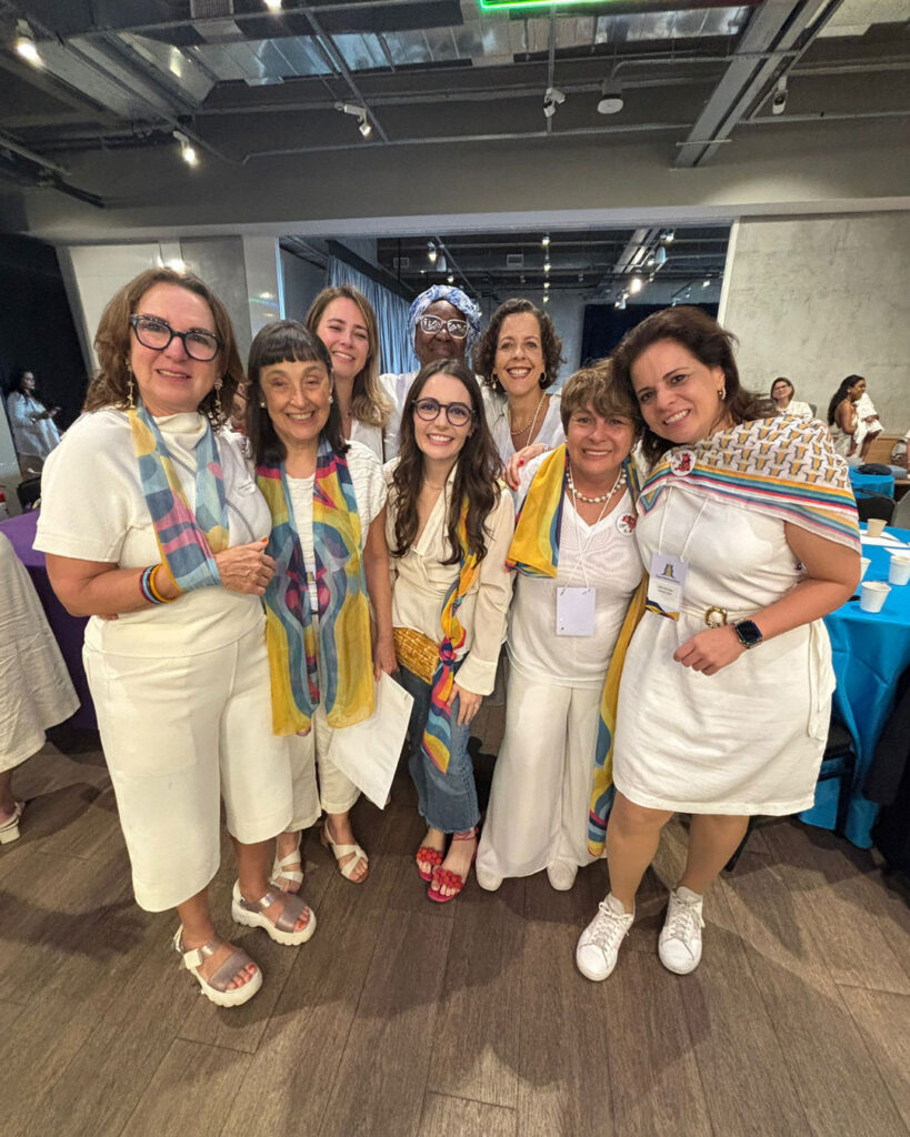 Grupo de mulheres reunidas em um evento, muitas vestidas de branco e usando lenços coloridos, posando para a foto.