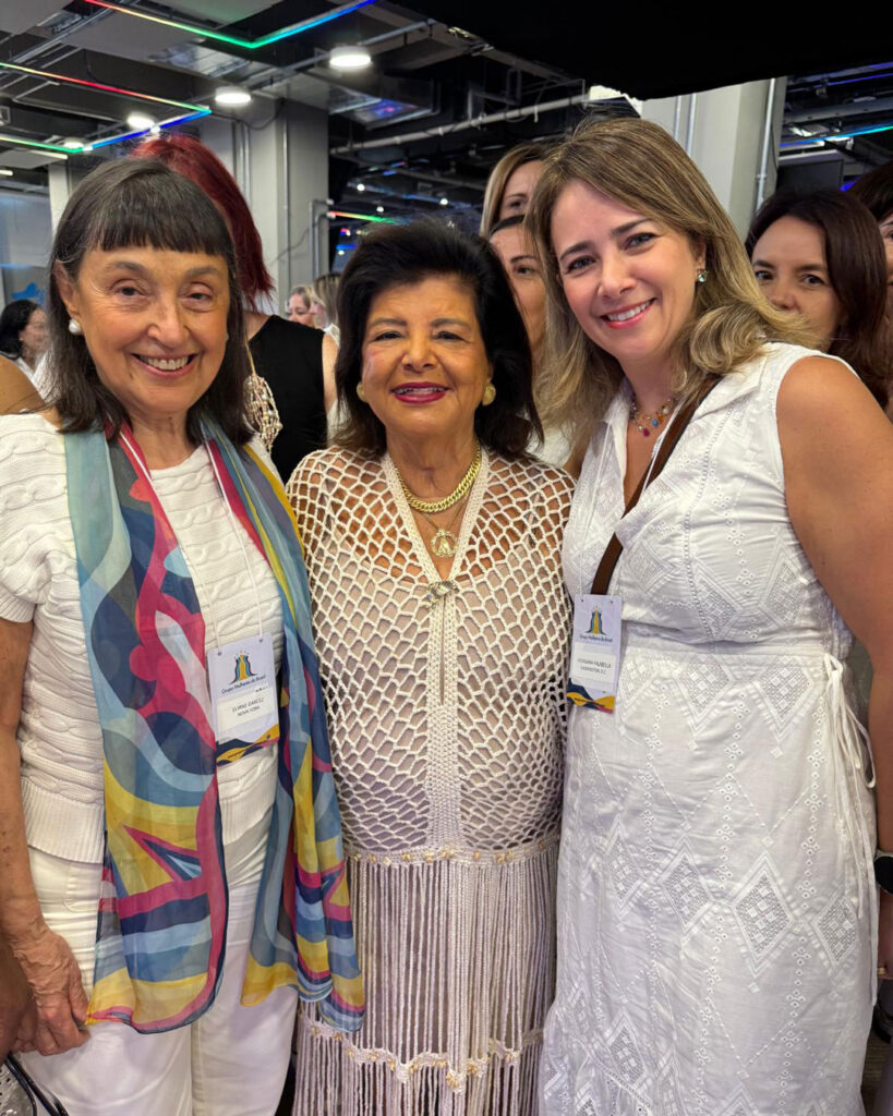 Três mulheres sorrindo lado a lado em um evento, posando para uma foto.